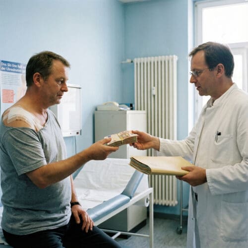 Ein Mann mit Schulterbandage übergibt einem Arzt im weißen Kittel ein dickes Geldbündel gegen ein umfangreiches Dokument.Ein Mann mit Schulterbandage übergibt einem Arzt im weißen Kittel ein dickes Geldbündel gegen ein umfangreiches Dokument.