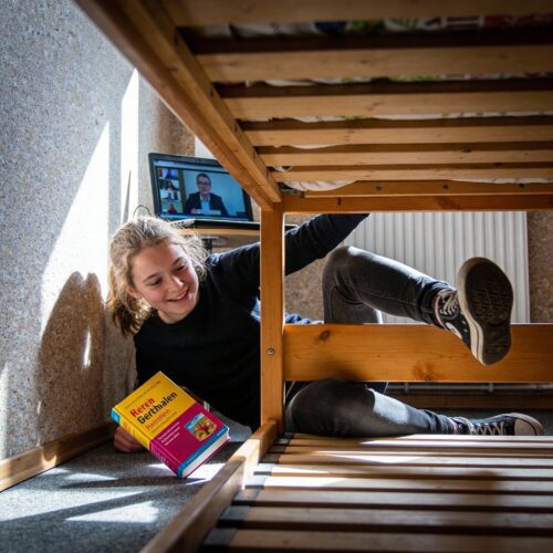 Eine Schülerin stolpert über die scharfe Kante eines Bettgestells, während sie nach einem Wörterbuch greift und der Online-Unterricht läuft.