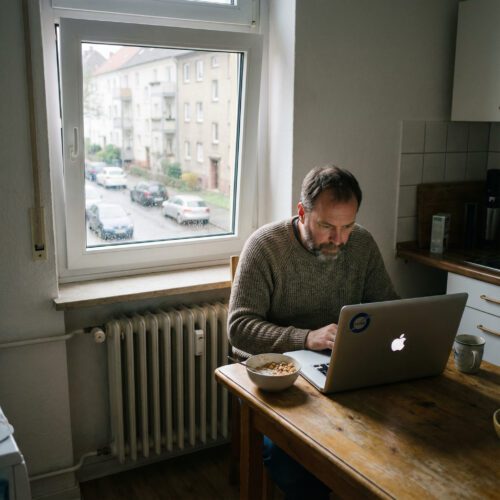 Mann arbeitet am Laptop mit ausländischem Firmenlogo an einem provisorischen Küchentisch neben einer Müslischale.